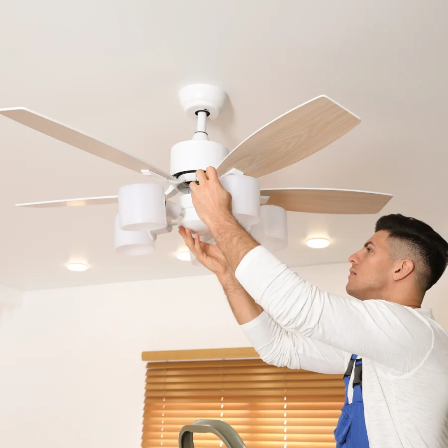 Electrician reaching up to install a white ceiling fan with light kit in a bright room