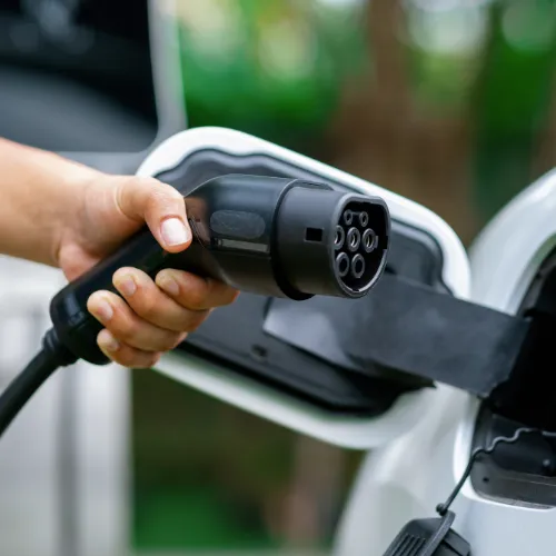Hand holding an EV charging connector next to the open charge port of a white electric vehicle