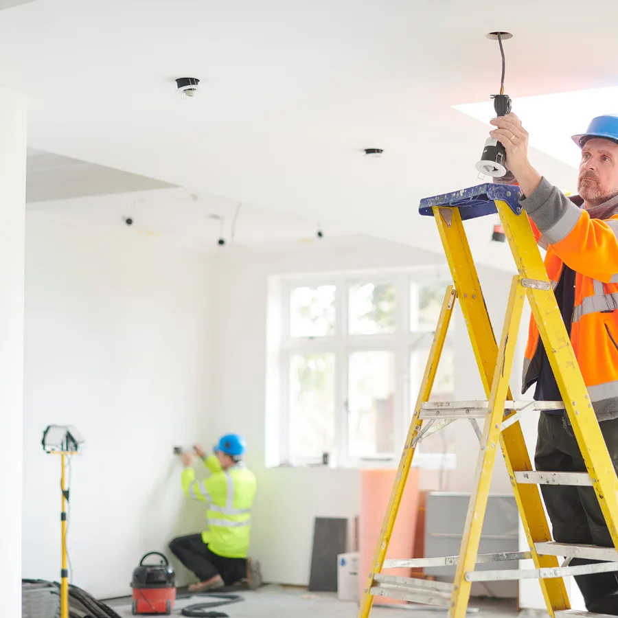 Electricians installing ceiling light fixtures and wall devices in a home under renovation