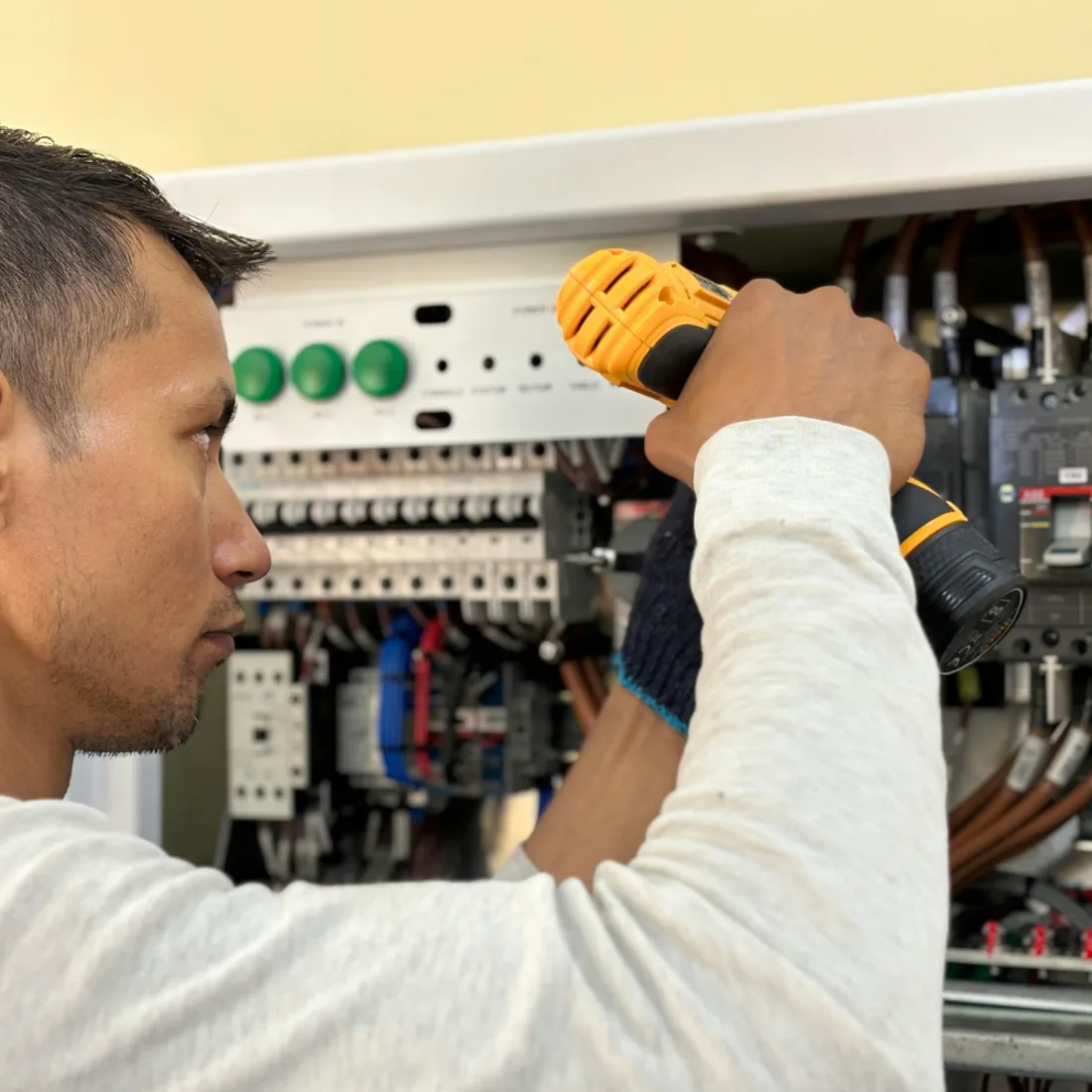 Electrician using a cordless drill while working inside an open electrical panel with rows of breakers