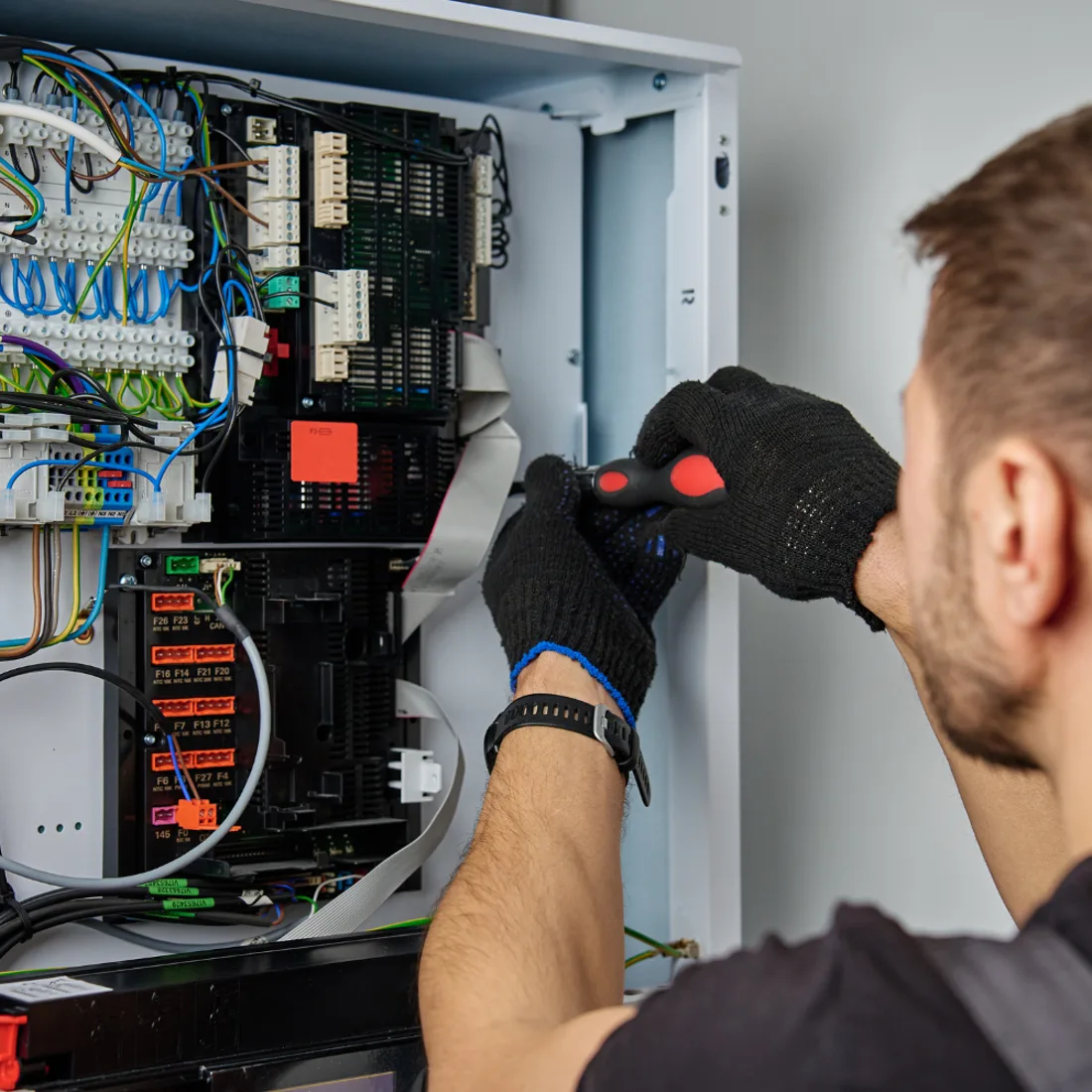Technician in gloves using a screwdriver inside an open electrical enclosure filled with wiring and terminals